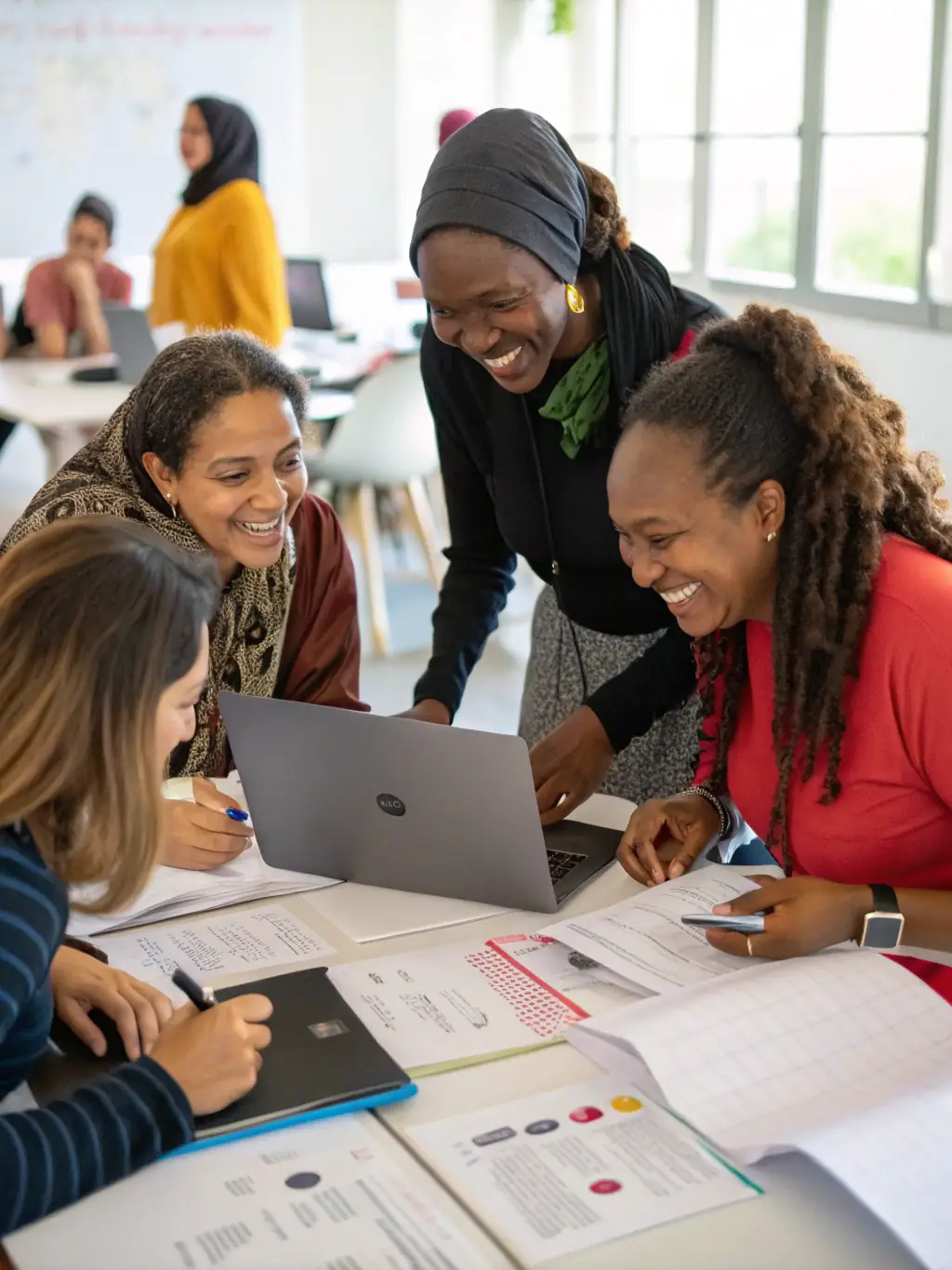 A group of women entrepreneurs actively participating in a hands-on workshop session, learning new business strategies and techniques, with laptops and notebooks spread across the table.